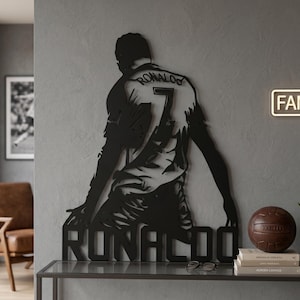 Op de afbeelding: Zwarte metalen wandkunst met een voetballer in knielende houding, met de naam "RONALDO" onderaan en het nummer "7" op het shirt. De naam van de speler staat ook op de achterkant van het shirt. Een decoratief stuk voor sportfans.