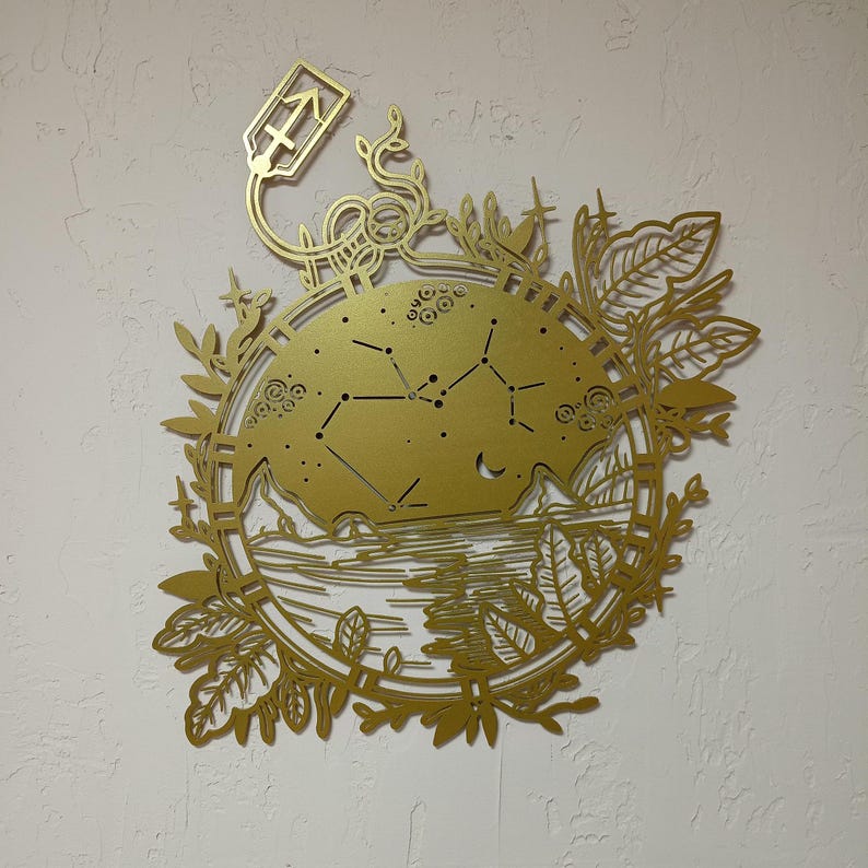 Puede incluir: Arte de pared de metal dorado con un dise&ntilde;o circular que presenta una constelaci&oacute;n, una luna creciente y una escena de paisaje. El dise&ntilde;o est&aacute; rodeado por un borde floral.
