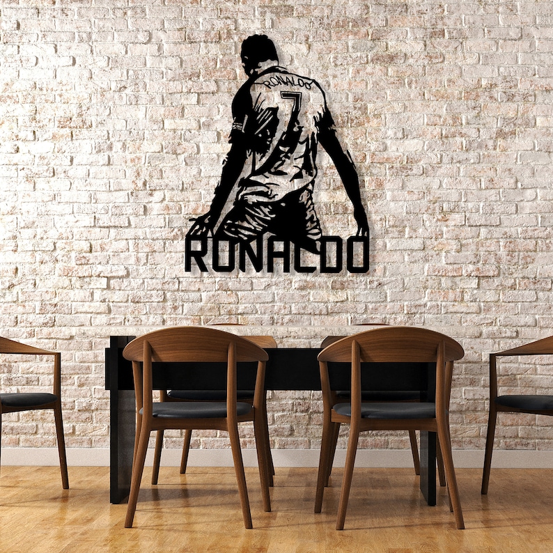 Op de afbeelding: Zwart metalen wandkunst van een voetballer in knielende pose, met de naam "RONALDO" eronder. De speler draagt een shirt met nummer 7. De kunst is gemonteerd op een witte bakstenen muur, boven een tafel en stoelen.