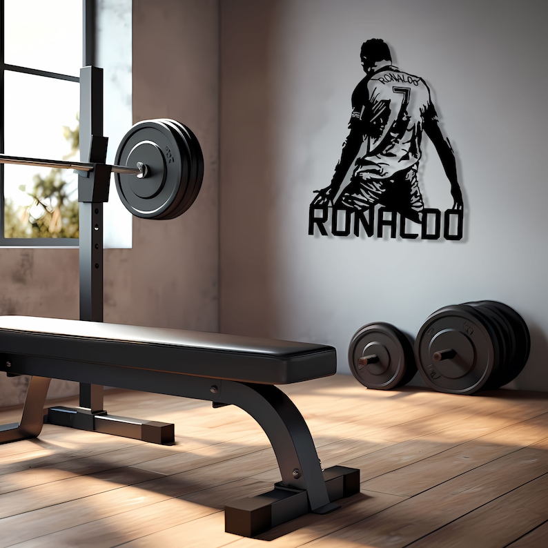 Op de afbeelding: Een zwarte fitnessruimte met een halterbank, halter met gewichten en dumbbells. Een zwart metalen wandkunstsilhouet van een voetballer met de naam "RONALDO" is aan de muur bevestigd. De kamer heeft een raam en houten vloeren.