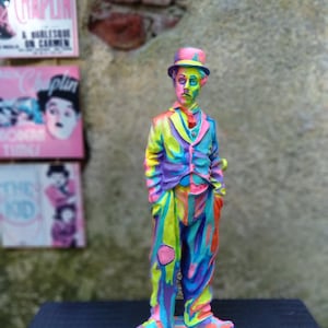 以下が含まれることがあります： ピンク、パープル、イエロー、ブルーの色のチャールズ・チャップリンのカラフルな置物。置物は黒い帽子とスーツを着ており、市松模様の台座の上に立っています。
