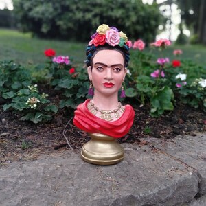 以下が含まれることがあります： 花冠と赤いショールをつけた、赤と金のフリーダ・カーロの胸像彫刻。彫刻は石の表面に置かれています。