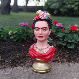 以下が含まれることがあります： 赤いショールと花冠をつけた、カラフルなセラミック製のフリーダ・カーロの胸像。胸像は金色の台座の上に置かれています。