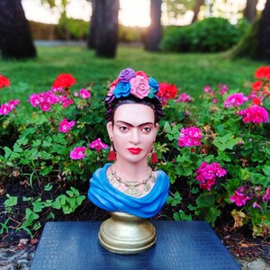 以下が含まれることがあります： 青い花冠をかぶった、青と金のフリア・カローの胸像。青いスカーフとイヤリングをしています。胸像は金の台座の上にあり、ピンクの花に囲まれています。