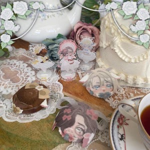 Może przedstawiać: Kolekcja akrylowych figurek i okrągłych przypinek z ilustracjami postaci w stylu anime w ubraniach z falbankami. Przedmioty są ułożone na drewnianej powierzchni obok białego czajnika i ciasta z lukrem.