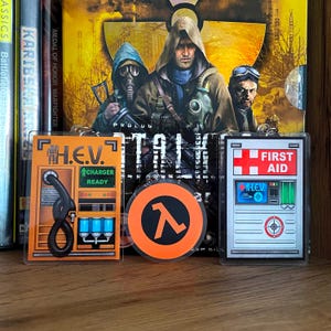 以下が含まれることがあります： ビデオゲームのイメージが描かれた3つのアクリルキーホルダー。左は長方形のオレンジ色のH.E.V.充電ステーション。中央は円形のオレンジ色のラムダ記号。右は赤い十字が描かれた長方形の銀色の応急処置ステーションです。