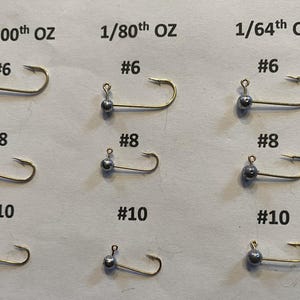 May include: Nine fishing jig heads with varying weights and hook sizes are displayed. The weights are labeled as 1/100th oz, 1/80th oz, and 1/64th oz. Hook sizes are #6, #8, and #10. Each jig head has a silver ball weight and a gold hook.