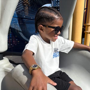 May include: A young person wearing a white t-shirt with blue text that says "Urban District 1972" and black shorts. They are wearing black sunglasses and a gold watch. The person is sitting on a white plastic slide.