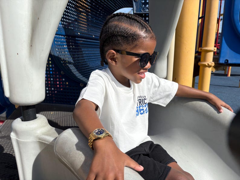 May include: A young person wearing a white t-shirt with blue text that says "Urban District 1972" and black shorts. They are wearing black sunglasses and a gold watch. The person is sitting on a white plastic slide.