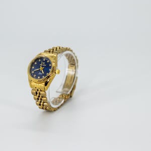 May include: A gold-tone wristwatch with a blue face and a gold-tone metal band. The watch has a round face with a gold-tone bezel and a gold-tone crown.