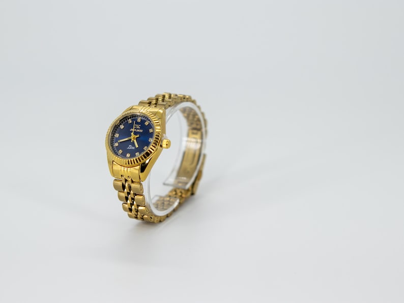 May include: A gold-tone wristwatch with a blue face and a gold-tone metal band. The watch has a round face with a gold-tone bezel and a gold-tone crown.