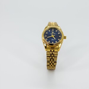 May include: A gold-tone wristwatch with a blue face and a gold metal band. The watch face has a white dial with black hour and minute hands, and a small second hand. The watch face also has a small date window at the 3 o'clock position. The watch band is a traditional link style.