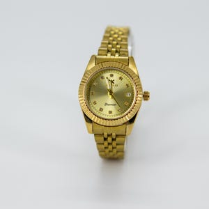 May include: A gold-toned wristwatch with a textured bezel and a matching link bracelet. The watch face is gold with diamond-like markers and the text "Brennan". The watch has a date display at the 3 o'clock position.