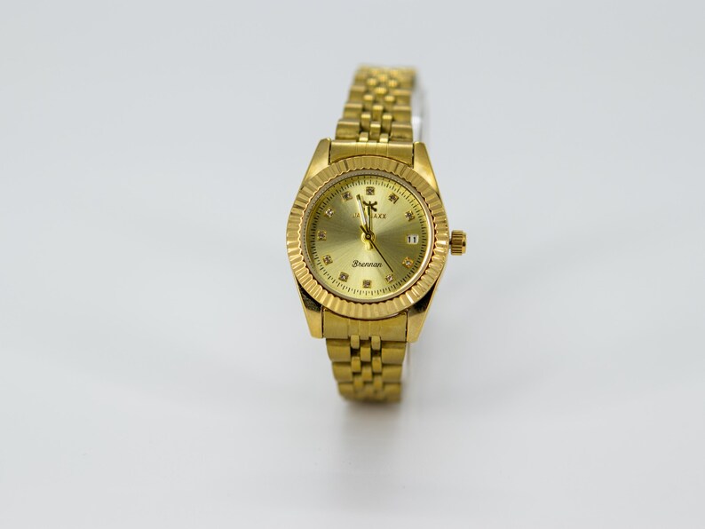 May include: A gold-toned wristwatch with a textured bezel and a matching link bracelet. The watch face is gold with diamond-like markers and the text "Brennan". The watch has a date display at the 3 o'clock position.