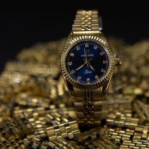 May include: A gold-tone wristwatch with a blue face and a gold metal band. The watch face has a gold-tone border with small diamonds around the edge. The watch face also has a gold-tone logo and the word "Isia" printed on it.