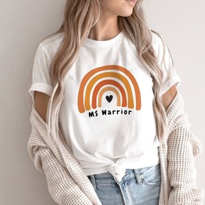 This model is wearing a white t-shirt that has a 4 striped rainbow. Each stripe is a different shade of orange. There is a small black heart in the center of the rainbow and below that in black text it says MS Warrior