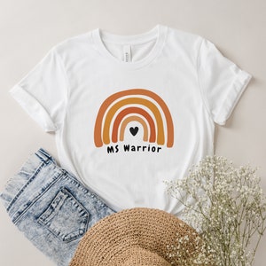 K&ouml;nnte beinhalten: Wei&szlig;es T-Shirt mit einem Regenbogen-Design und dem Text "MS Warrior" mit einem schwarzen Herzen in der Mitte des Regenbogens.