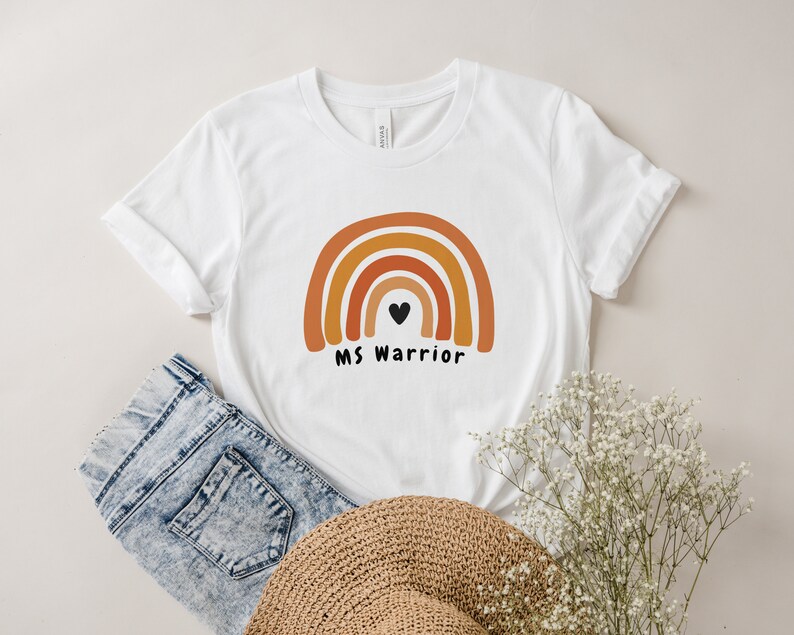 K&ouml;nnte beinhalten: Wei&szlig;es T-Shirt mit einem Regenbogen-Design und dem Text "MS Warrior" mit einem schwarzen Herzen in der Mitte des Regenbogens.