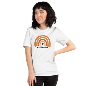 K&ouml;nnte beinhalten: Wei&szlig;es T-Shirt mit einem Regenbogen-Design und dem Text "MS Warrior" mit einem kleinen schwarzen Herzen in der Mitte des Regenbogens.