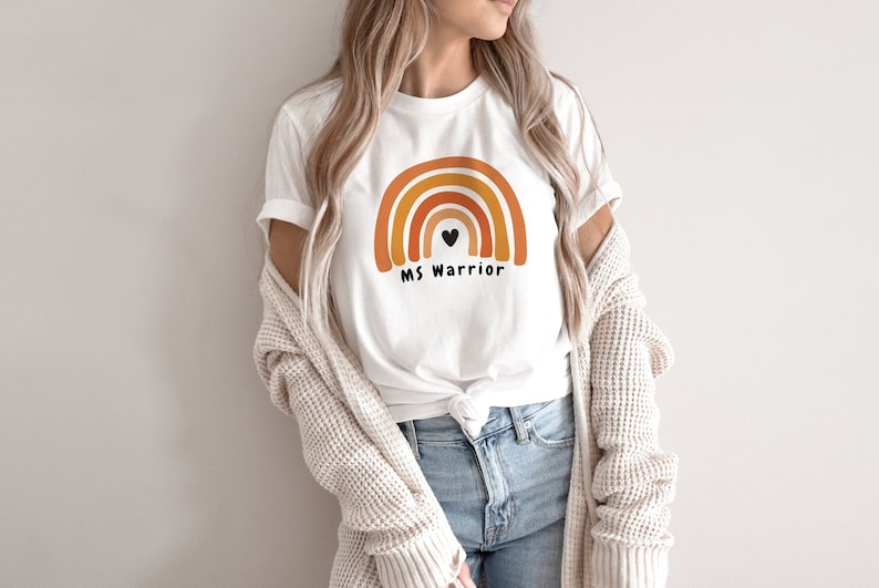 This model is wearing a white t-shirt that has a 4 striped rainbow. Each stripe is a different shade of orange. There is a small black heart in the center of the rainbow and below that in black text it says MS Warrior