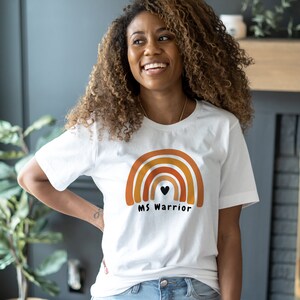 K&ouml;nnte beinhalten: Wei&szlig;es T-Shirt mit einem Regenbogen-Motiv und dem Text "MS Warrior" in Schwarz. Der Regenbogen ist in Orange-, Gelb- und Rott&ouml;nen gehalten.