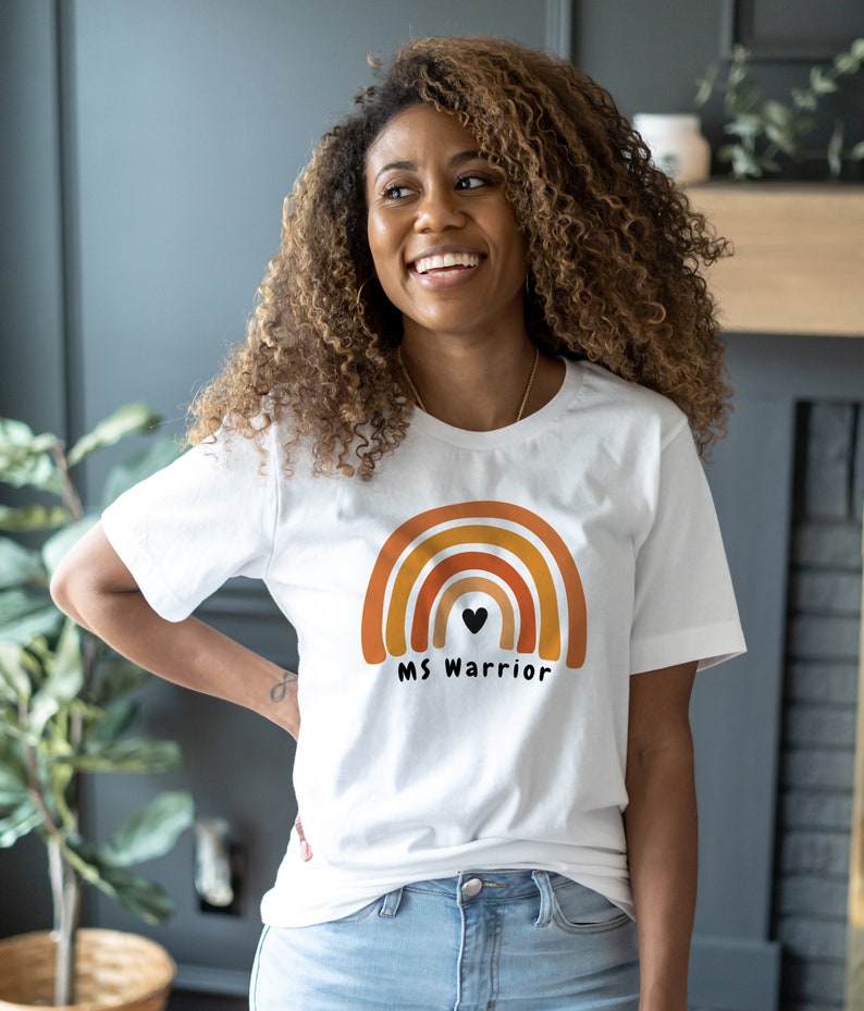 K&ouml;nnte beinhalten: Wei&szlig;es T-Shirt mit einem Regenbogen-Motiv und dem Text "MS Warrior" in Schwarz. Der Regenbogen ist in Orange-, Gelb- und Rott&ouml;nen gehalten.