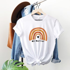 K&ouml;nnte beinhalten: Wei&szlig;es T-Shirt mit einem Regenbogen-Design und dem Text "MS Warrior" mit einem kleinen schwarzen Herzen in der Mitte des Regenbogens.