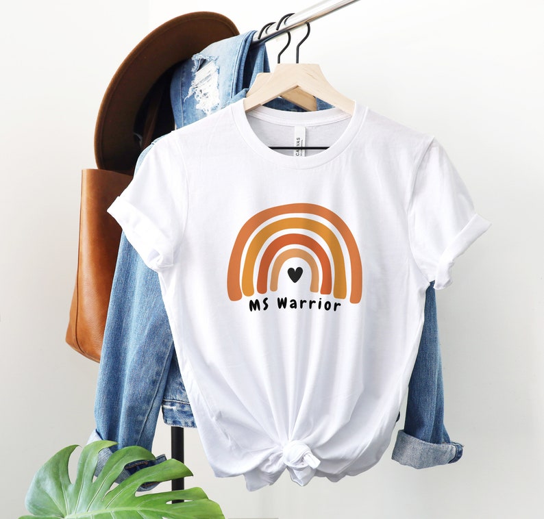 K&ouml;nnte beinhalten: Wei&szlig;es T-Shirt mit einem Regenbogen-Design und dem Text "MS Warrior" mit einem kleinen schwarzen Herzen in der Mitte des Regenbogens.