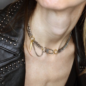 Può includere: Una collana a catena d'argento e oro con un piccolo ciondolo d'argento e un anello d'oro. La collana è indossata da una persona che indossa una giacca di pelle nera.
