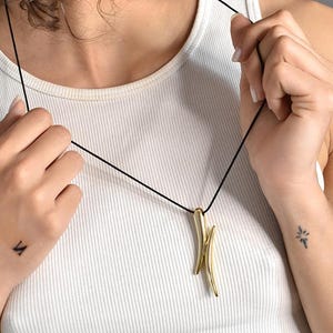 May include: A gold necklace with a pendant featuring two curved, pointed shapes. The necklace is on a black cord and is being worn by a person.
