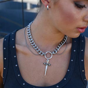 May include: Silver-toned necklace featuring a thick chain and a dagger pendant. The necklace has a layered design with a smaller chain and a ring detail. The model is wearing a dark gray tank top with silver studs.