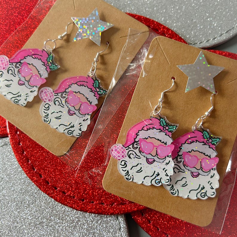 May include: Two pairs of Santa Claus earrings with pink hats and sunglasses, and a star-shaped top. The earrings are on brown cardboard backing, with silver fish hook ear wires. The Santa faces have white beards and pink cheeks.