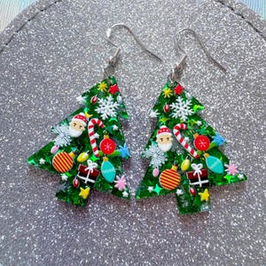 May include: A pair of Christmas tree-shaped earrings. The green earrings are decorated with festive ornaments, including Santa faces, candy canes, snowflakes, and wrapped gifts. The earrings have silver-colored hooks.