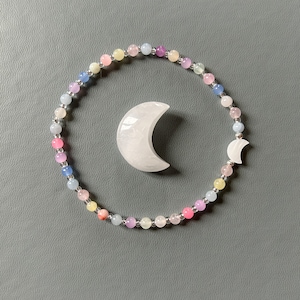 May include: A colorful beaded bracelet with a crescent moon charm and a separate moon-shaped stone. The bracelet features small, round beads in pastel colors, including pink, blue, yellow, and purple. The moon-shaped elements are white.