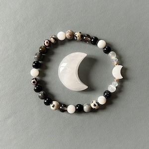 May include: A bracelet made of black, white and grey stones with a white crescent moon charm. The bracelet is on a grey background.