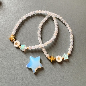 May include: Two beaded bracelets with clear beads and gold star accents. Each bracelet features a gold star charm, a white disc with a star, and a light blue star charm. A light blue star-shaped pendant is also present.