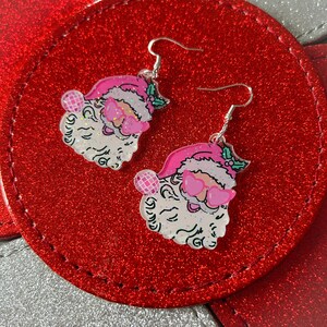 May include: A pair of festive Santa Claus earrings. Each earring features a Santa face with a pink hat, heart-shaped sunglasses, and a glittery beard. The earrings are suspended from silver-colored fish hook ear wires. The background is a red, glittery surface.