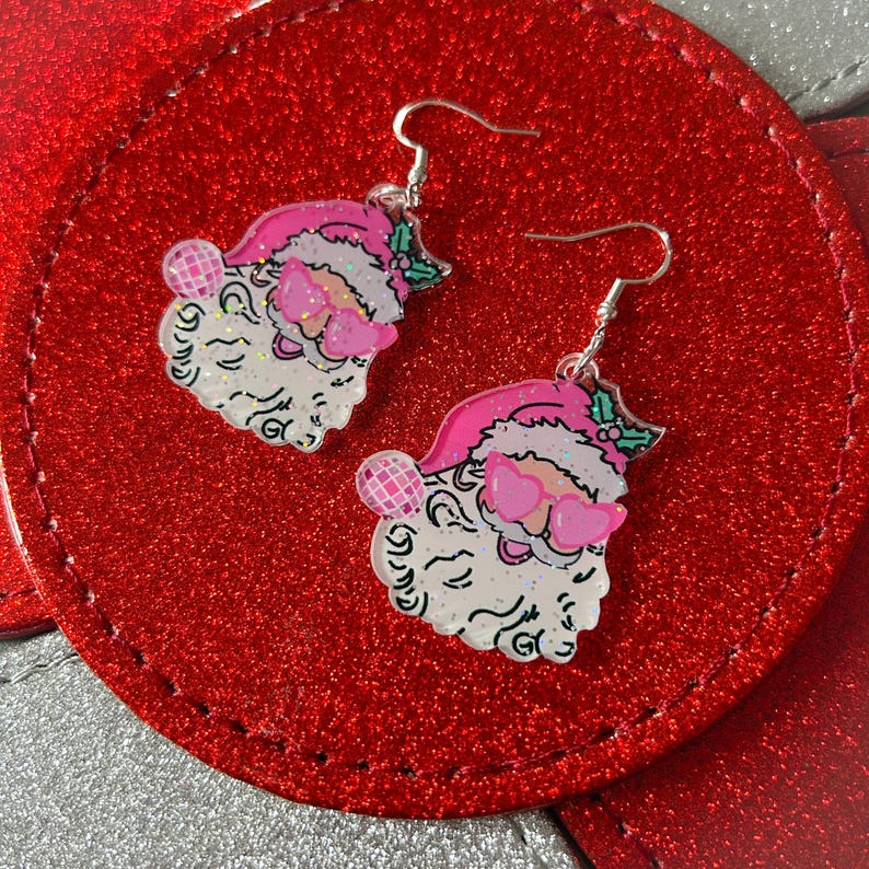 May include: A pair of festive Santa Claus earrings. Each earring features a Santa face with a pink hat, heart-shaped sunglasses, and a glittery beard. The earrings are suspended from silver-colored fish hook ear wires. The background is a red, glittery surface.