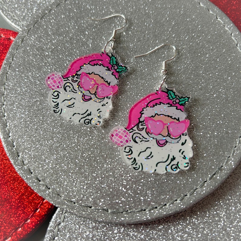 May include: A pair of festive Santa Claus earrings. Each earring features a Santa face with a pink hat, heart-shaped sunglasses, and a white beard. The earrings have silver-colored hooks and are set against a silver background.