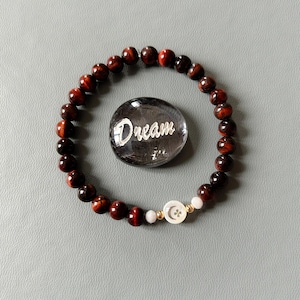 May include: Red tiger's eye beaded bracelet with a white shell charm featuring a crescent moon and flower design. The bracelet is accompanied by a small glass stone with the word "Dream" engraved on it.