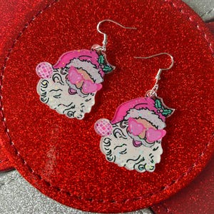 May include: A pair of Christmas-themed earrings featuring Santa Claus faces with pink sunglasses and hats. The earrings have silver hooks and are set against a red, glittery background. Each Santa face has a white beard and a pink disco ball.