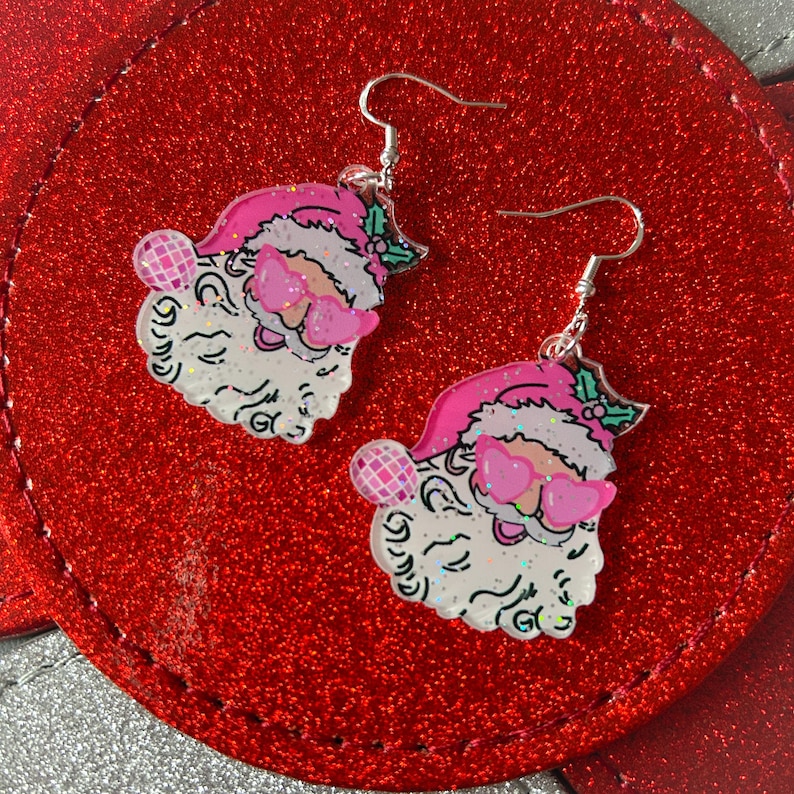 May include: A pair of Christmas-themed earrings featuring Santa Claus faces with pink sunglasses and hats. The earrings have silver hooks and are set against a red, glittery background. Each Santa face has a white beard and a pink disco ball.