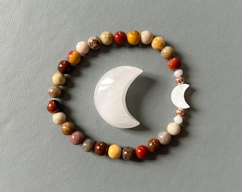 Può includere: Un braccialetto di perline con pietre dai toni della terra, tra cui rosso, marrone e giallo. Un ciondolo a forma di mezzaluna bianca e un piccolo charm a forma di luna sono anch'essi presenti. Il braccialetto e i ciondoli sono esposti su una superficie grigia.