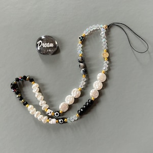 May include: A white and black beaded phone charm with a clear crystal and gold accents. The charm also includes a small black stone with a white eye design. The charm is attached to a black string.