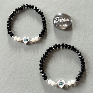 May include: Two black beaded bracelets with white heart charms and blue evil eye details. The bracelets are on a gray surface with a small glass stone that says "Dream".