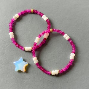 May include: Two pink and white beaded bracelets with gold accents. The bracelets are stacked on top of each other. A small, iridescent star-shaped stone is in the foreground.