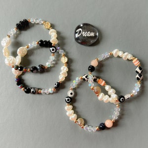 May include: Three beaded bracelets featuring white pearls, black and white agate beads, and crystal beads. The bracelets have gold accents, evil eye charms, and a seashell charm. A gray stone with the word "Dream" written on it is also in the image.