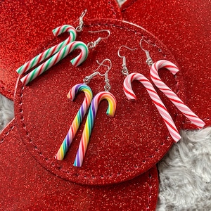 May include: A collection of candy cane earrings in various colors, including red and white stripes, green and white stripes, and rainbow hues. The earrings are displayed on a red, glittery surface, suggesting a festive theme.