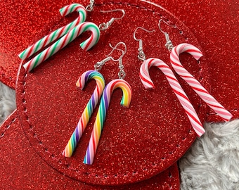 May include: A collection of candy cane earrings in various colors, including red and white stripes, green and white stripes, and rainbow hues. The earrings are displayed on a red, glittery surface, suggesting a festive theme.