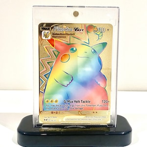 Puede incluir: Una tarjeta Pokémon Pikachu VMAX dorada con colores arcoíris, exhibida en una funda acrílica transparente sobre un soporte negro. La tarjeta presenta el texto "Pikachu VMAX" y "G-Max Volt Tackle".